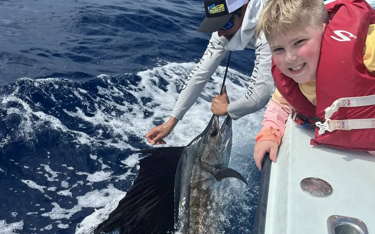 Young angler beside a sailfish released boatside off Miami with the skyline behind - Nomad Fishing Charters